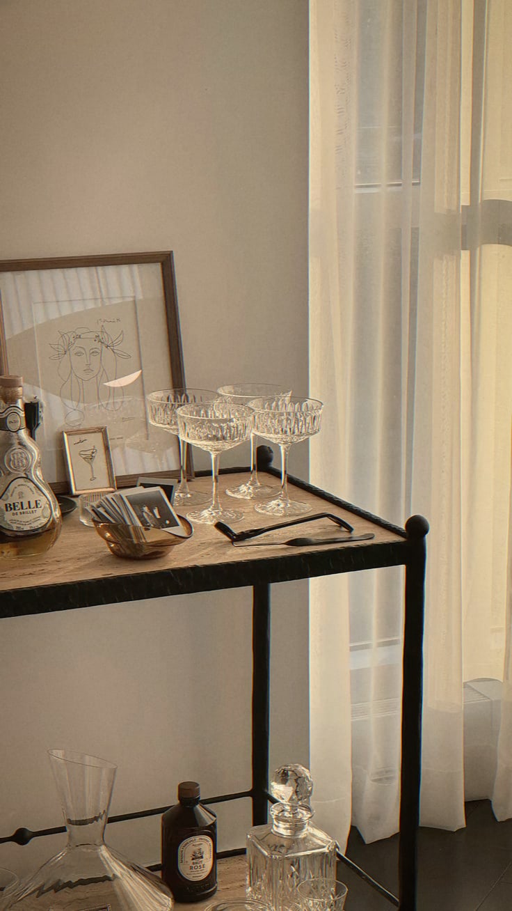 Bar cart with crystal coupes, a decanter and a framed line drawing by a sheer curtain
